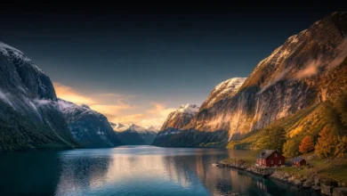 Stunning aerial view of a majestic Norwegian fjord landscape with deep blue water, steep mountain cliffs, and traditional red wooden cabins under a golden sunset reflecting Nerwey geography.