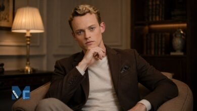 A sophisticated portrait of British actor Anson Boon sitting in a study, wearing a brown blazer over a beige sweater.
