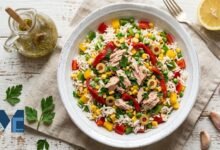 A colorful bowl of authentic Spanish ensalada de arroz featuring fluffy white rice, tuna, green olives, corn, peas, and roasted red peppers on a rustic table.