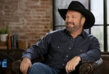 Garth Brooks smiling in a casual pose wearing a black cowboy hat and dark shirt in a modern indoor setting.
