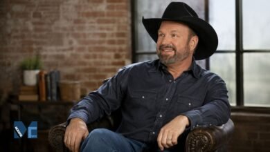 Garth Brooks smiling in a casual pose wearing a black cowboy hat and dark shirt in a modern indoor setting.