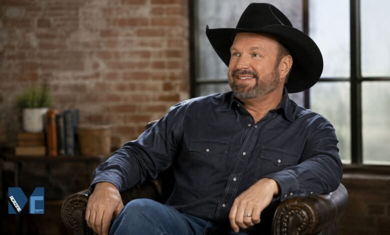 Garth Brooks smiling in a casual pose wearing a black cowboy hat and dark shirt in a modern indoor setting.