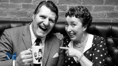 A black and white vintage photograph of British comedian Tommy Cooper laughing with a mug in his hand, while his wife Gwen Cooper smiles and playfully points at the mug.