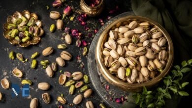 A premium flat-lay arrangement of fresh pistachios, both in-shell and shelled with vibrant green kernels, displayed in vintage brass bowls on a dark slate surface with scattered dried rose petals.