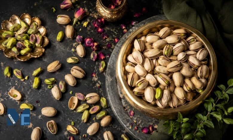 A premium flat-lay arrangement of fresh pistachios, both in-shell and shelled with vibrant green kernels, displayed in vintage brass bowls on a dark slate surface with scattered dried rose petals.