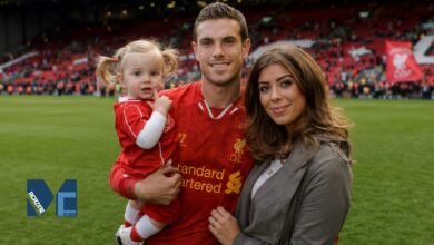 Rebecca Burnett smiling alongside her husband, Liverpool footballer Jordan Henderson, who is holding their young daughter on the Anfield pitch during a stadium lap of honor.