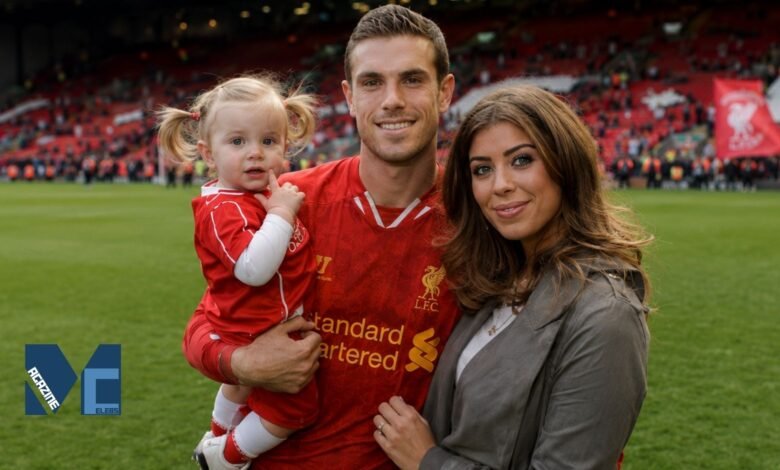 Rebecca Burnett smiling alongside her husband, Liverpool footballer Jordan Henderson, who is holding their young daughter on the Anfield pitch during a stadium lap of honor.