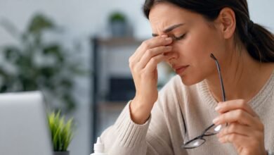 Woman experiencing dry eye symptoms while working on a laptop with eye drops and glasses on a desk in a modern workspace.